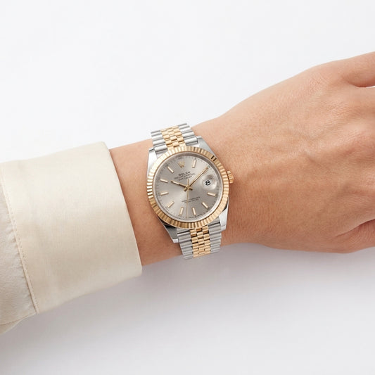 A two-tone stainless steel and gold wristwatch with a silver dial, worn on a person's wrist. The watch features a fluted gold-tone bezel and a two-tone link bracelet, with a cream-colored dress shirt cuff visible on the left.