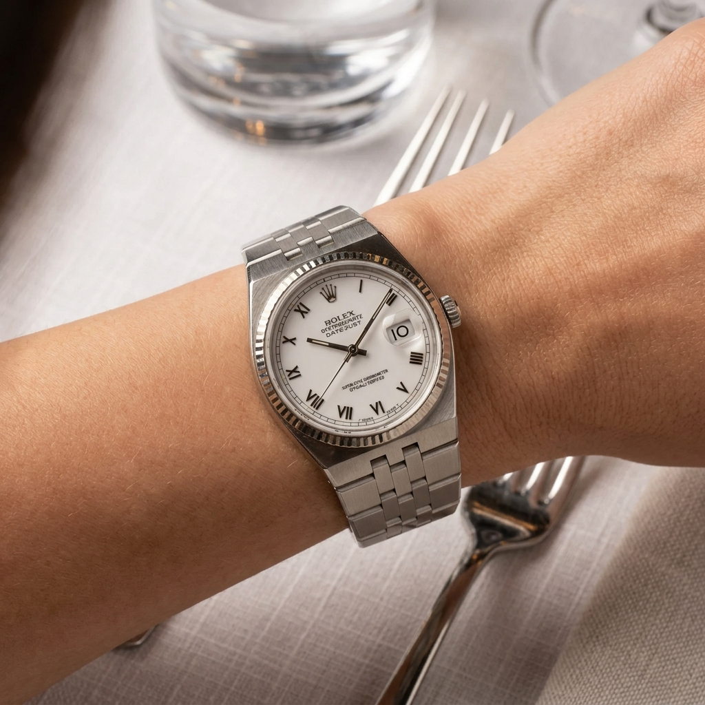 A Rolex Datejust watch with a silver stainless steel bracelet and white dial, worn on a person's wrist over a light-colored tablecloth with silverware and a glass in the background.