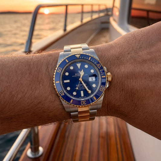 A Rolex Submariner watch with a blue dial and blue bezel, paired with a two-tone steel and gold bracelet, worn on a person's wrist on a wooden boat deck at sunset.