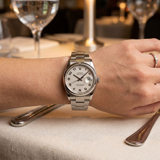 A silver stainless steel wristwatch with a white dial and date window, worn on a person's wrist in a restaurant setting with blurred wine glasses and tableware in the background.