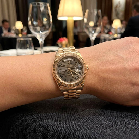 A gold-tone Rolex Day Date watch with a textured dark dial and date window, shown worn on a person's wrist in a dimly lit restaurant setting with wine glasses and table lamps in the background.