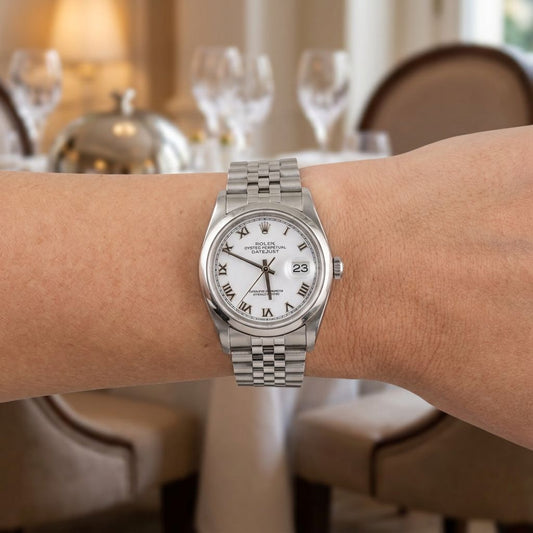 A Rolex wristwatch with a silver-tone metal bracelet and white dial, worn on a person's wrist in a softly blurred indoor dining setting.
