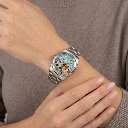 A silver stainless steel watch with a colorful floral dial is shown on a person's wrist, wearing a taupe knit sweater.