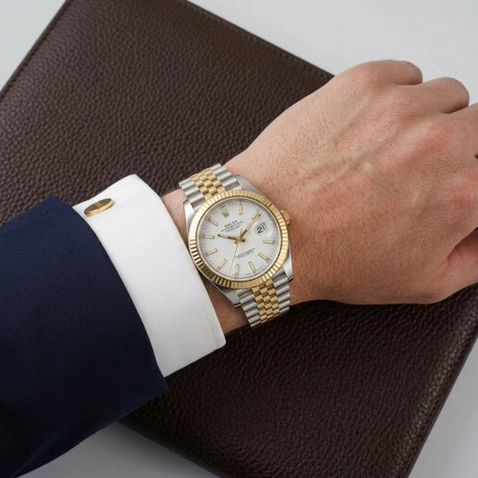 A Rolex Datejust watch with a two-tone gold and silver steel bracelet and a white dial, shown on a person's wrist wearing a white dress shirt and dark navy suit jacket, resting on a brown textured leather surface.