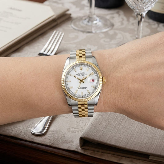 A Rolex Datejust watch with a two-tone silver and gold stainless steel bracelet, gold-tone fluted bezel, and white dial, shown worn on a person's wrist in a dining setting.