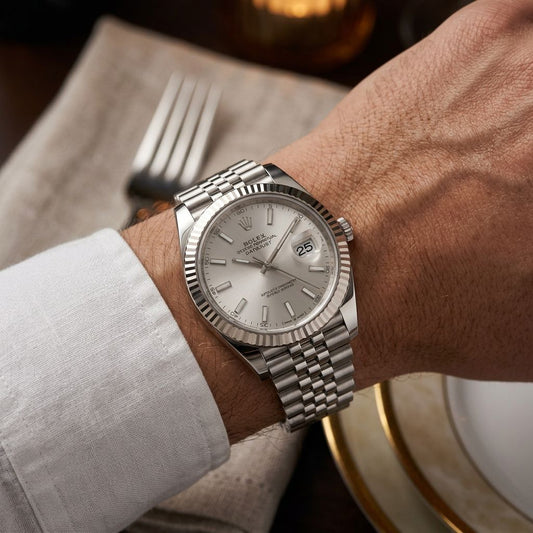 A silver Rolex Datejust watch with a stainless steel Oyster bracelet, worn on a person's wrist over a white dress shirt cuff, with a fork and plate blurred in the background.