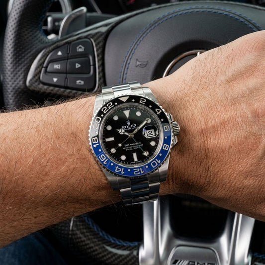 A Rolex Submariner Date watch with a black dial and blue bezel, worn on a person's wrist in front of a carbon-fiber car steering wheel.