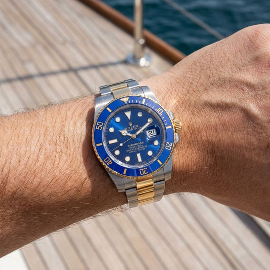 A Rolex Submariner Date watch with a blue dial and blue bezel, featuring a two-tone silver and gold stainless steel case and bracelet, worn on a person's wrist on a wooden deck.