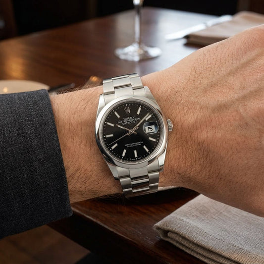 A stainless steel Rolex Oyster Perpetual watch with a black dial and date window, worn on a person's wrist over a dark gray dress shirt cuff, set against a blurred wooden table background.