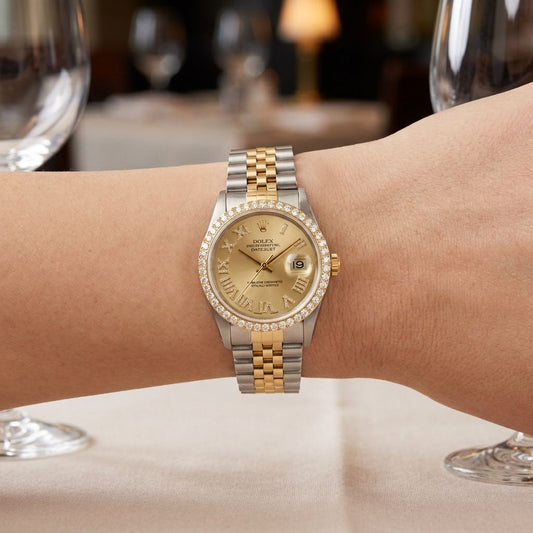 A Rolex Datejust watch with a two-tone gold and silver steel bracelet, a diamond-encrusted bezel, and a champagne dial, shown worn on a wrist in a blurred restaurant setting.
