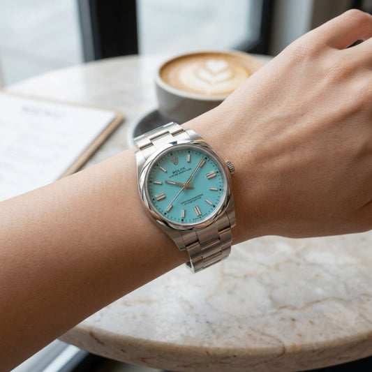 A silver stainless steel wristwatch with a light blue dial and matching blue hour markers, worn on a person's wrist in a cafe setting with a latte in the background.
