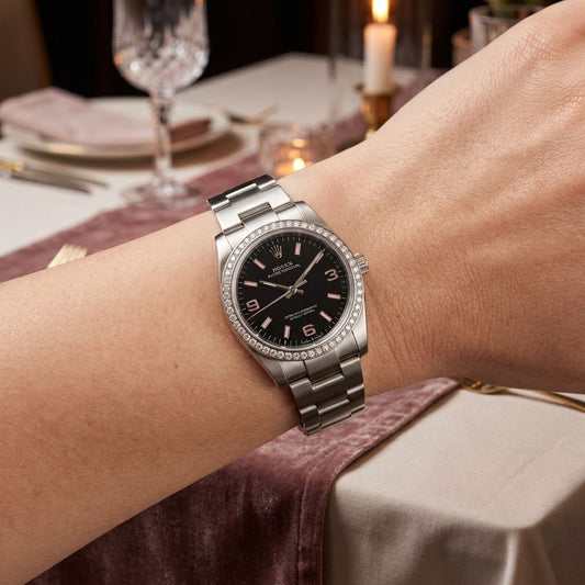 A silver stainless steel wristwatch with a black dial and diamond-accented bezel, worn on a person's wrist in a elegantly lit dining setting.