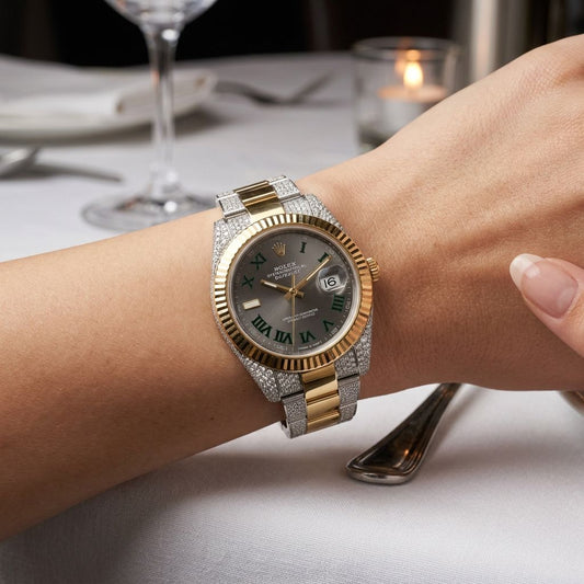 A luxury two-tone watch with a gold and silver-tone case, diamond-encrusted bezel and bracelet, and a dark gray dial with green Roman numerals, shown worn on a person's wrist in a dining setting.