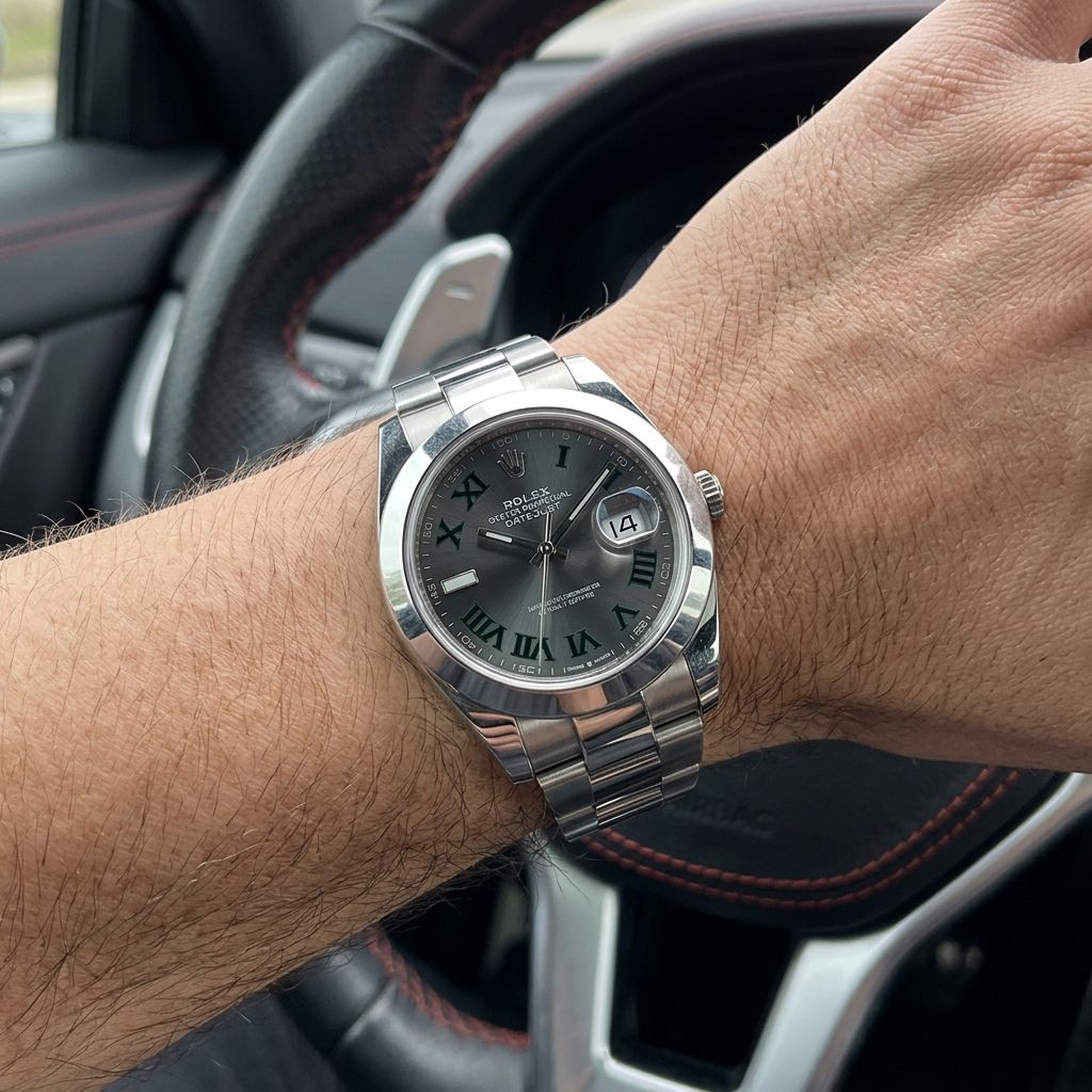 A silver stainless steel wristwatch with a dark gray dial and Roman numeral hour markers, worn on a person's wrist inside a car with a leather steering wheel visible in the background.