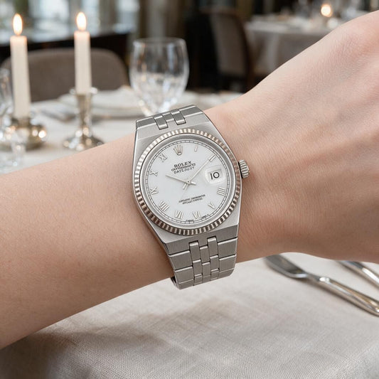A silver Rolex Datejust watch with a white dial and fluted bezel, worn on a person's wrist in a softly blurred restaurant setting with lit candles and tableware in the background.